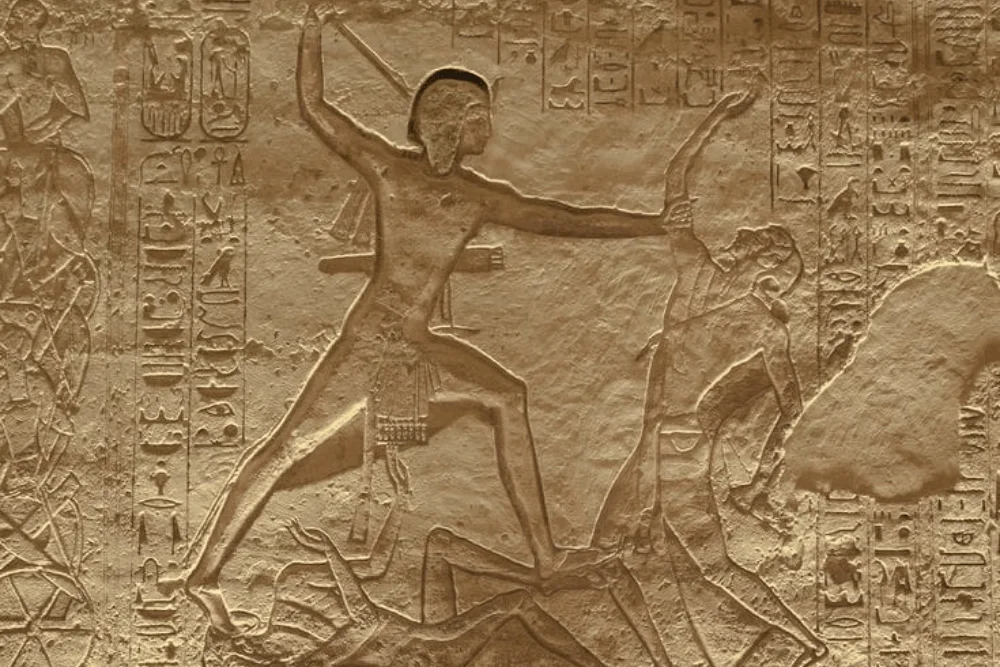 Ancient Egyptian relief showing Ramesses II in battle, striking an enemy, surrounded by hieroglyphs.